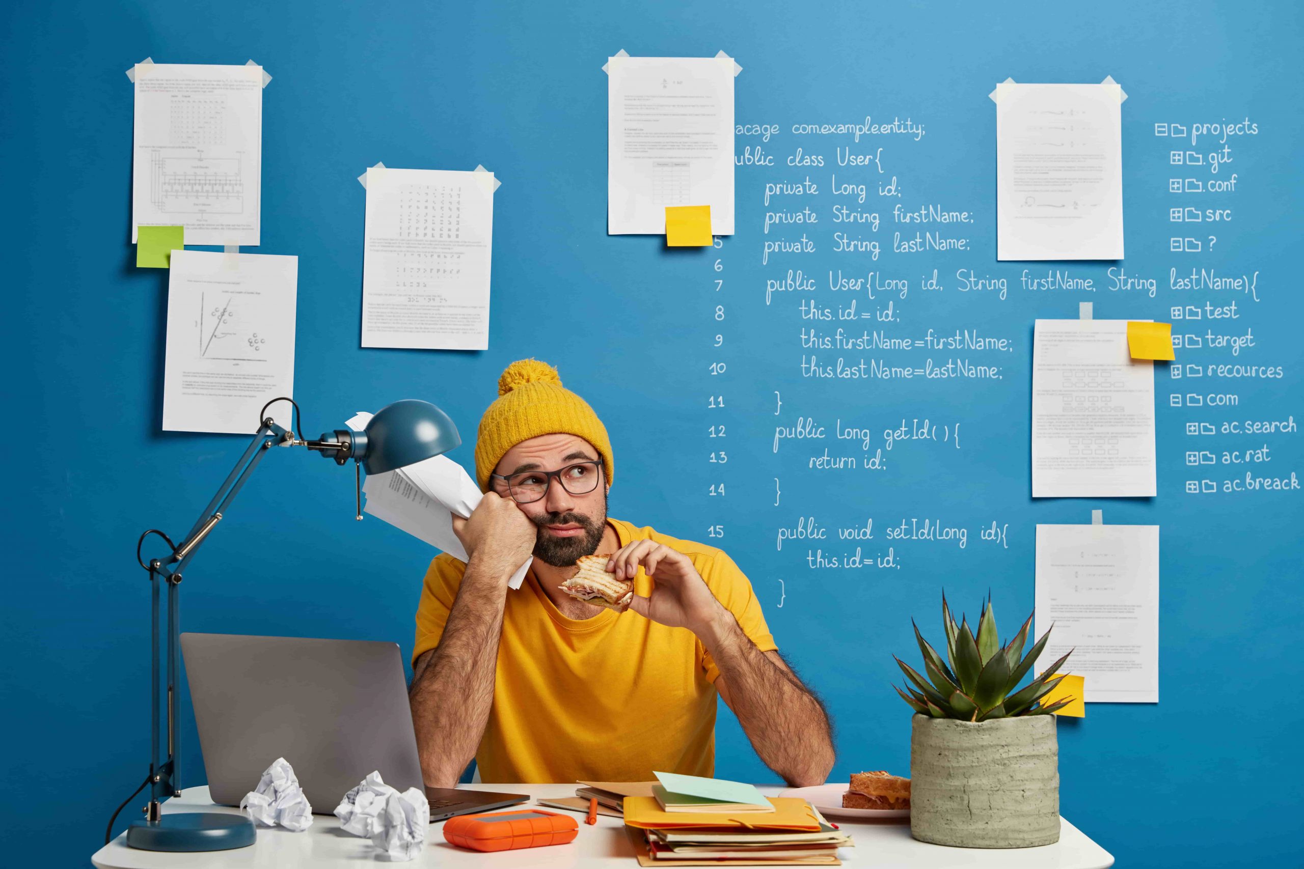 thoughtful-male-programmer-software-developer-ponders-program-code-looks-away-eats-burger-holds-papers-wears-yellow-clothes-spends-time-making-project-min